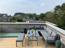 Modern rooftop terrace with gray outdoor furniture, table set with drinks, and small pool, surrounded by trees and buildings.