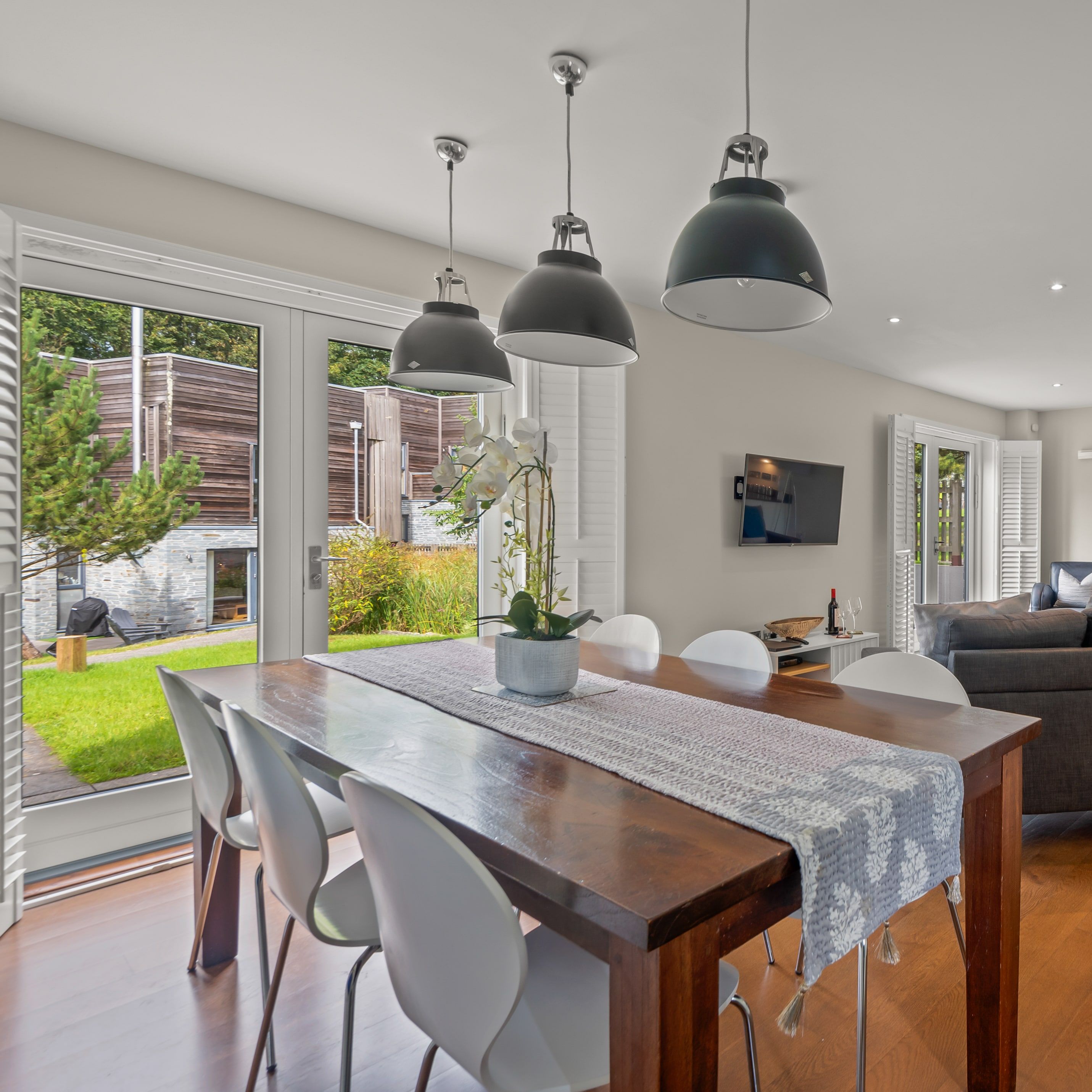 Modern open-plan living and dining area with wooden floors, a large wooden dining table with white chairs, gray sofas, pendant lighting, and glass doors opening to a garden.