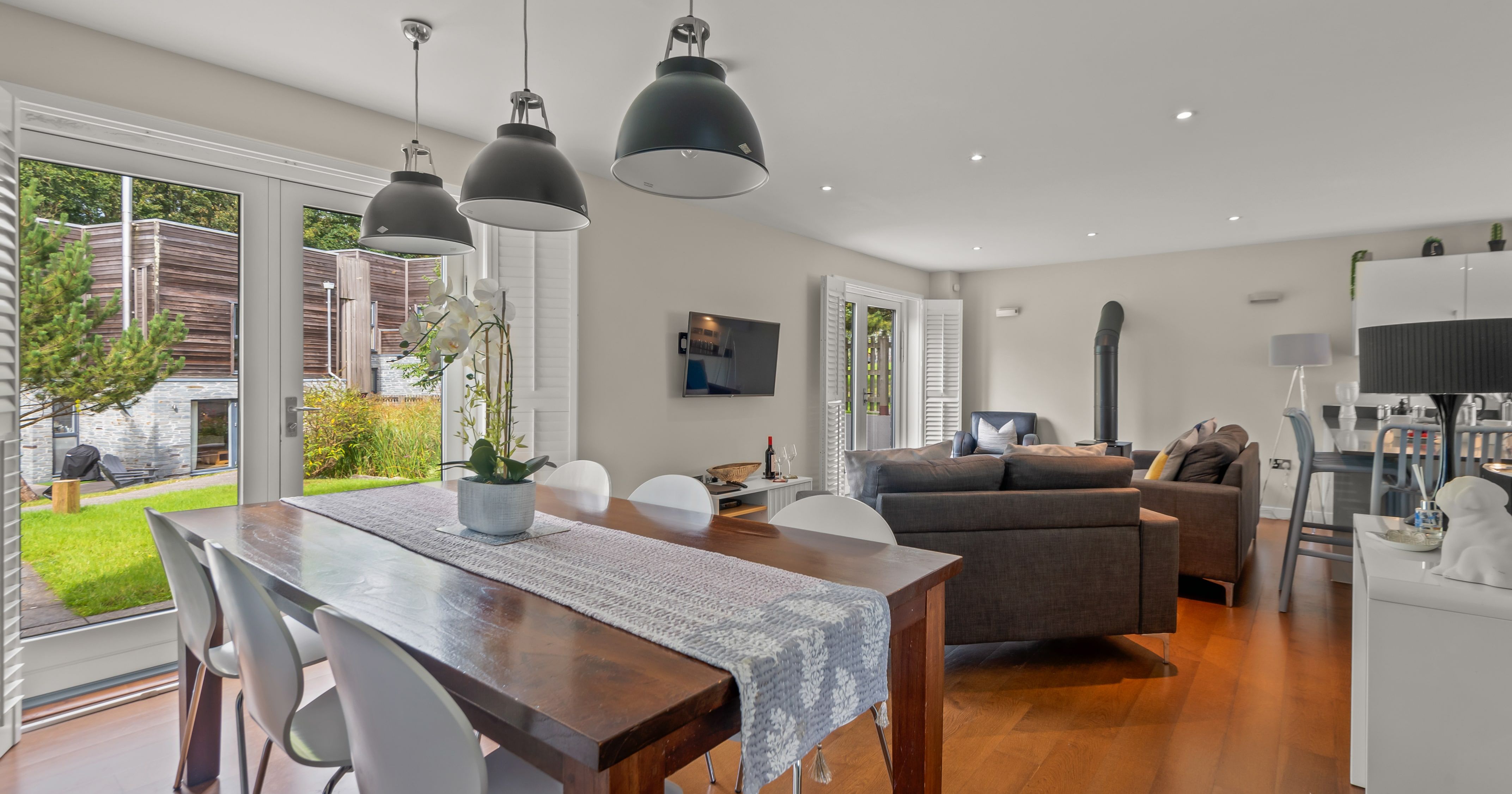 Modern open-plan living and dining area with wooden floors, a large wooden dining table with white chairs, gray sofas, pendant lighting, and glass doors opening to a garden.