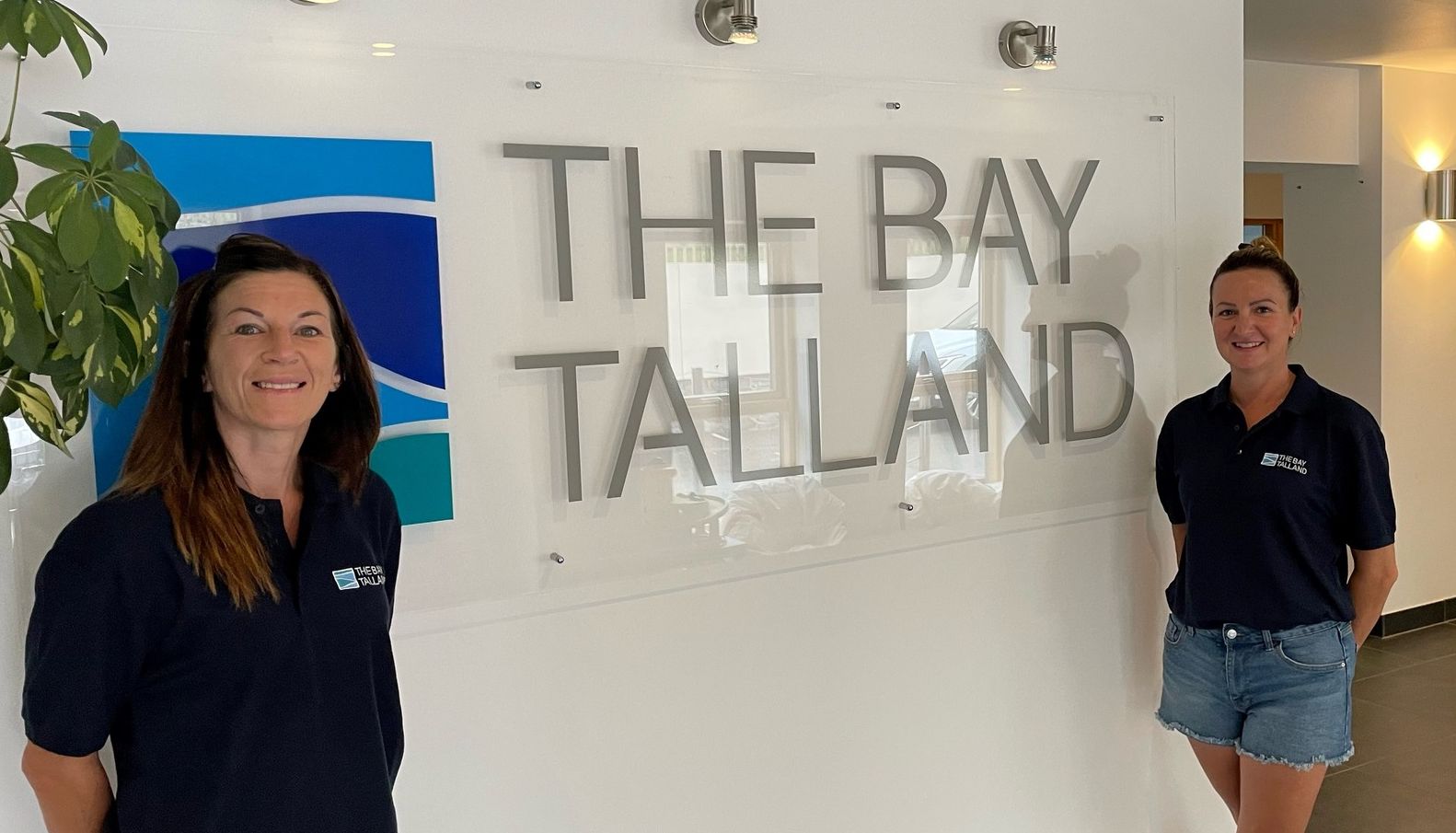 Two women standing in front of a sign that says 'The Bay Talland' inside a modern building.