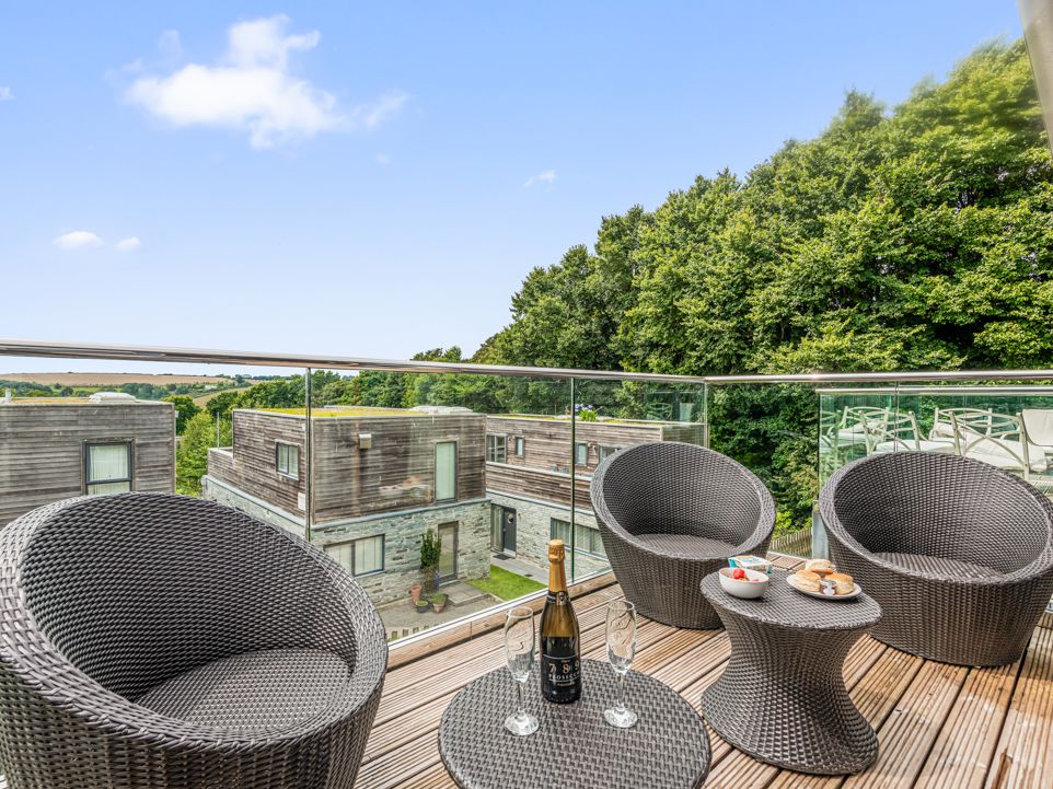 Modern outdoor balcony with wicker chairs, glass railing, snacks, and drinks overlooking contemporary residential buildings and trees.
