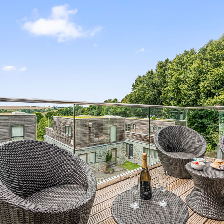 Modern outdoor balcony with wicker chairs, glass railing, snacks, and drinks overlooking contemporary residential buildings and trees.