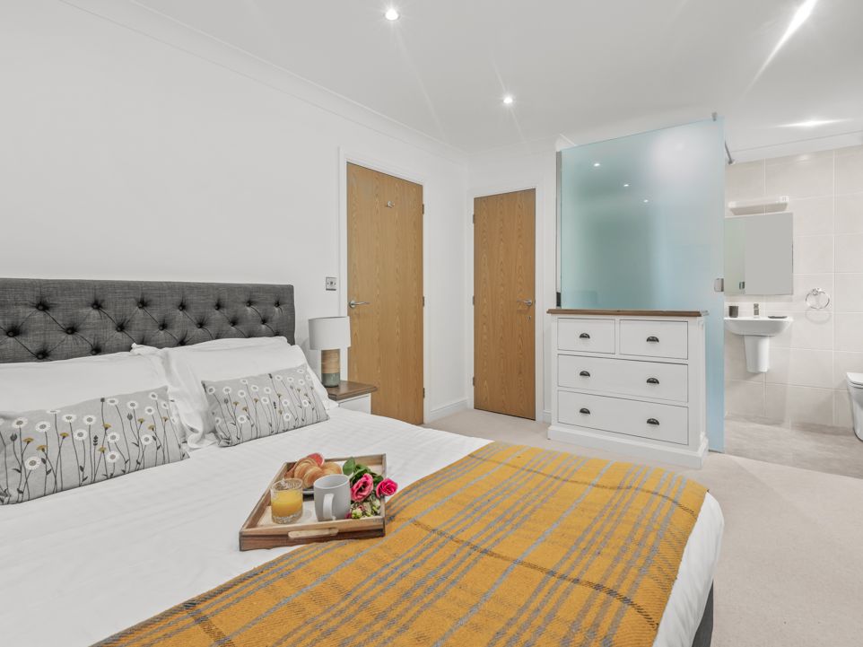 Modern bedroom with a king-sized bed, decorative pillows, and a breakfast tray, featuring a dresser and a partial view of an en-suite bathroom.