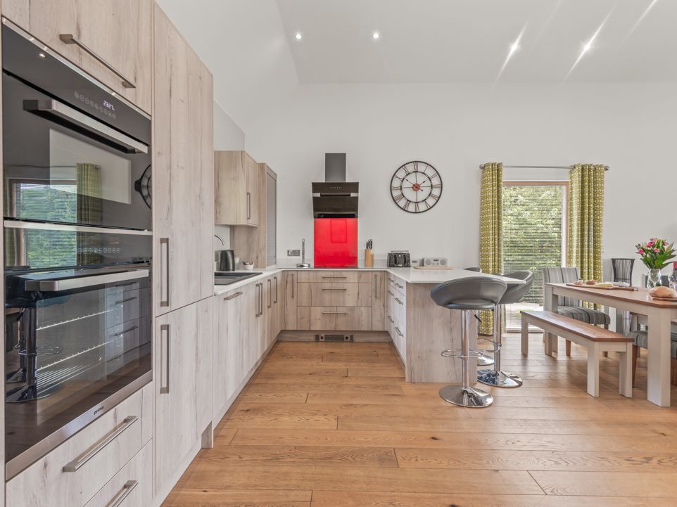 Modern kitchen and dining area with wooden cabinets, built-in ovens, a red backsplash, bar stools, and a dining table with flowers.
