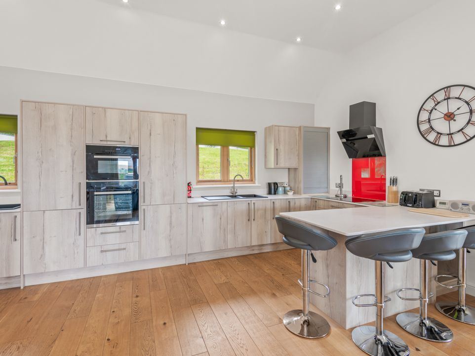 Modern kitchen interior with light wood cabinets, island with bar stools, and large wall clock.