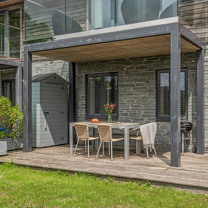 Outdoor patio area with a dining table, chairs, a lounge chair, and a shed against a stone house wall.