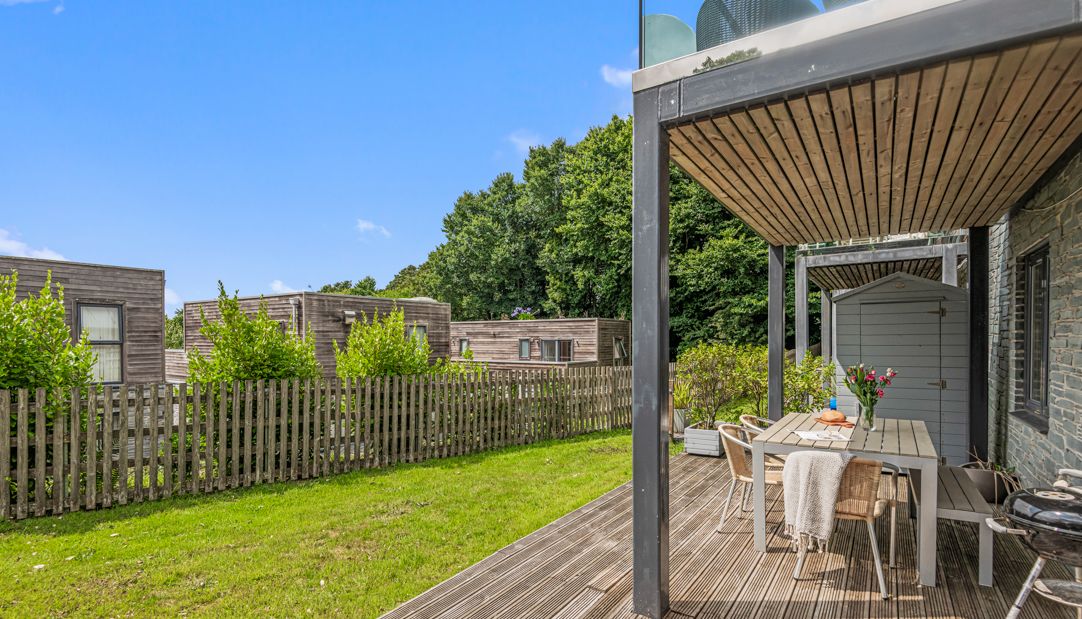 Modern wooden patio with outdoor dining set next to green lawn and wooden fence, surrounded by trees and contemporary houses.
