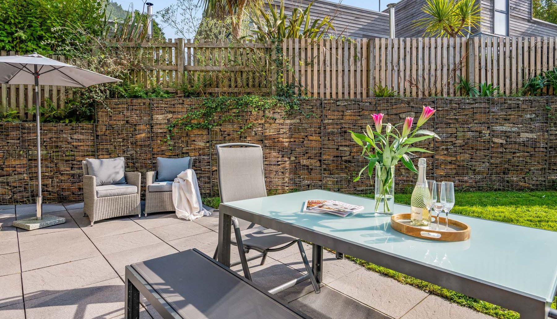 Modern patio with outdoor seating, a glass table with flowers, drinks, and a magazine, surrounded by greenery and wooden fencing.