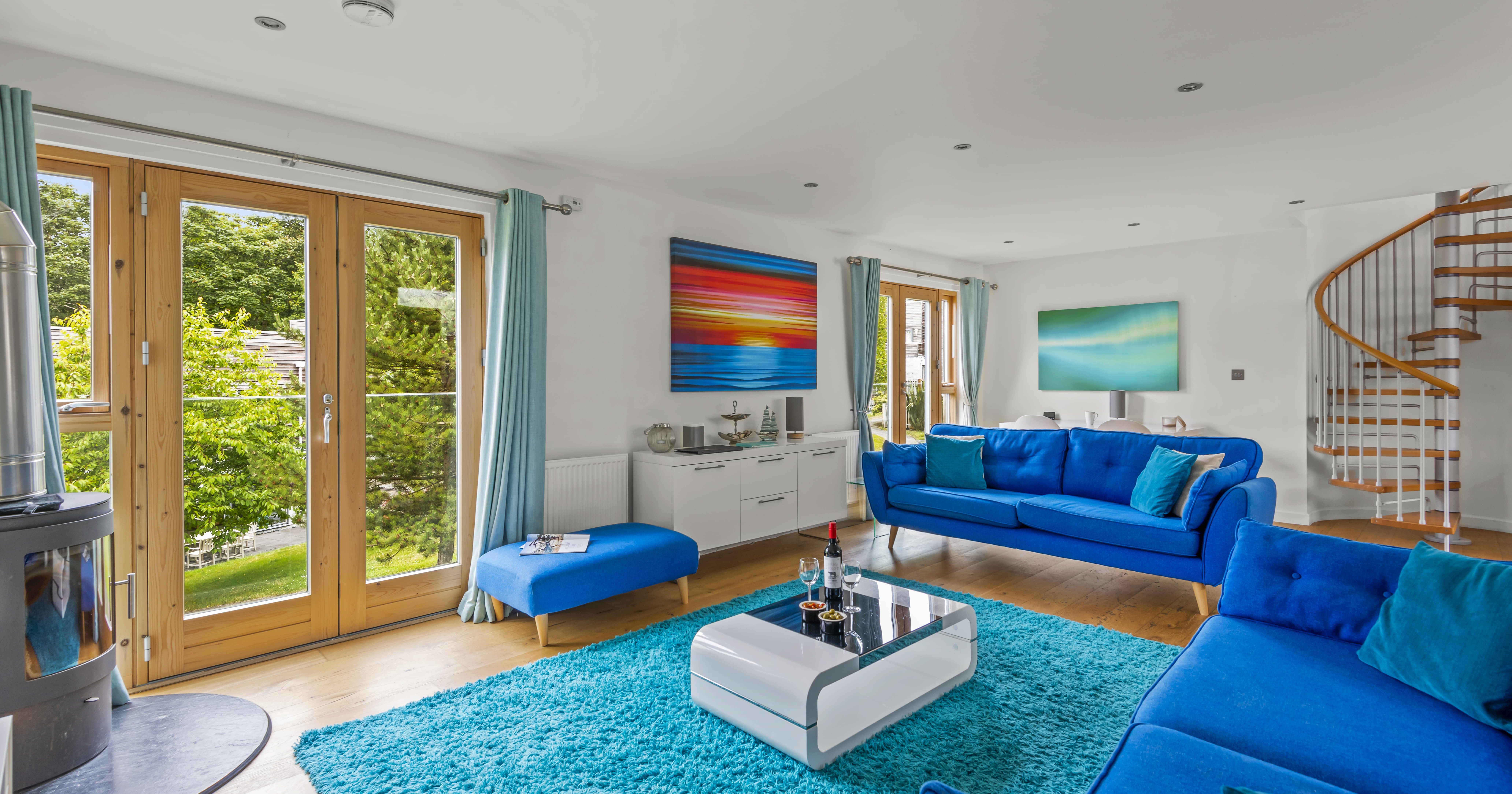 Modern living room with bright blue sofas, a turquoise rug, glass coffee table, large windows with wooden frames, contemporary artwork, and a spiral staircase.