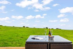 Outdoor table with glasses, wine, book, and snacks overlooking a green field under a blue sky with clouds.