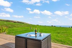 Outdoor patio table with a bottle of champagne, two glasses, an open book, and a bowl of fruit on a deck overlooking a large grassy field and distant ocean view.