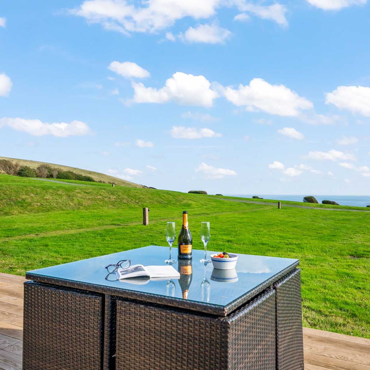Outdoor patio table with a bottle of champagne, two glasses, an open book, and a bowl of fruit on a deck overlooking a large grassy field and distant ocean view.