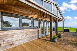 Modern wooden house with large windows and deck overlooking a grassy landscape and blue sky.