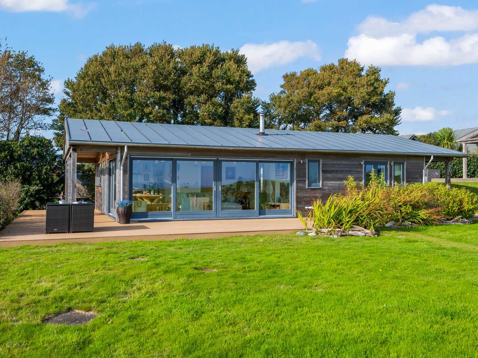Modern single-story house with large glass windows and a spacious deck, surrounded by green lawn and trees.