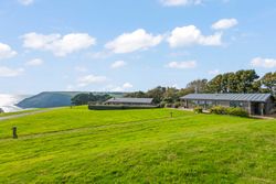 Modern houses with large windows on a grassy hill overlooking the sea and coastal cliffs under a blue sky with scattered clouds.
