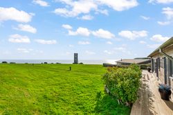 Coastal house with a large green lawn overlooking the sea under a bright blue sky with scattered clouds.