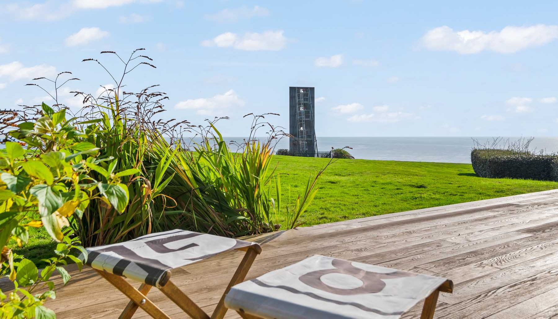 Coastal view from a wooden deck with two chairs, grassy lawn, plants, and a modern tower overlooking the sea.