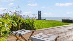 Coastal view from a wooden deck with two chairs, grassy lawn, plants, and a modern tower overlooking the sea.