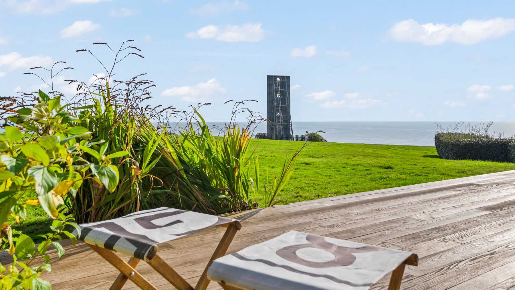 Coastal view from a wooden deck with two chairs, grassy lawn, plants, and a modern tower overlooking the sea.