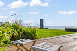Coastal view from a wooden deck with two chairs, grassy lawn, plants, and a modern tower overlooking the sea.