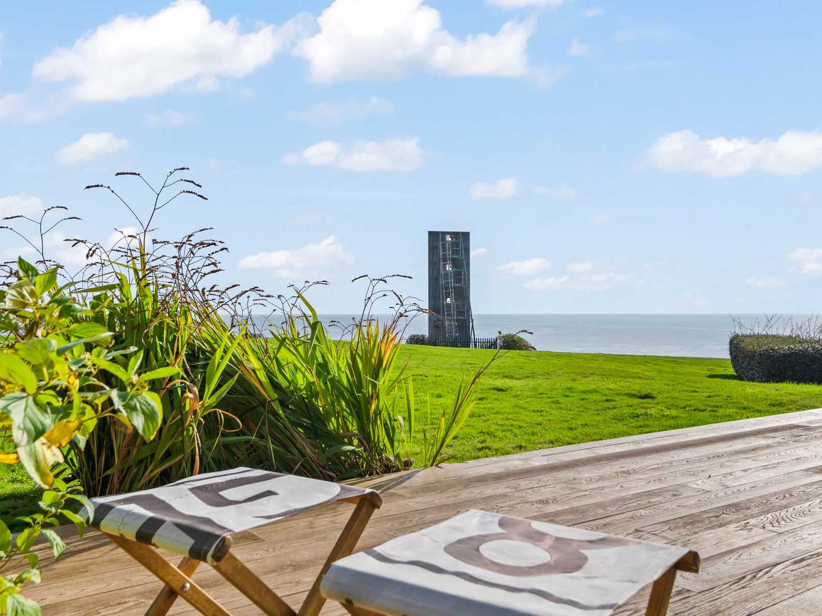 Coastal view from a wooden deck with two chairs, grassy lawn, plants, and a modern tower overlooking the sea.