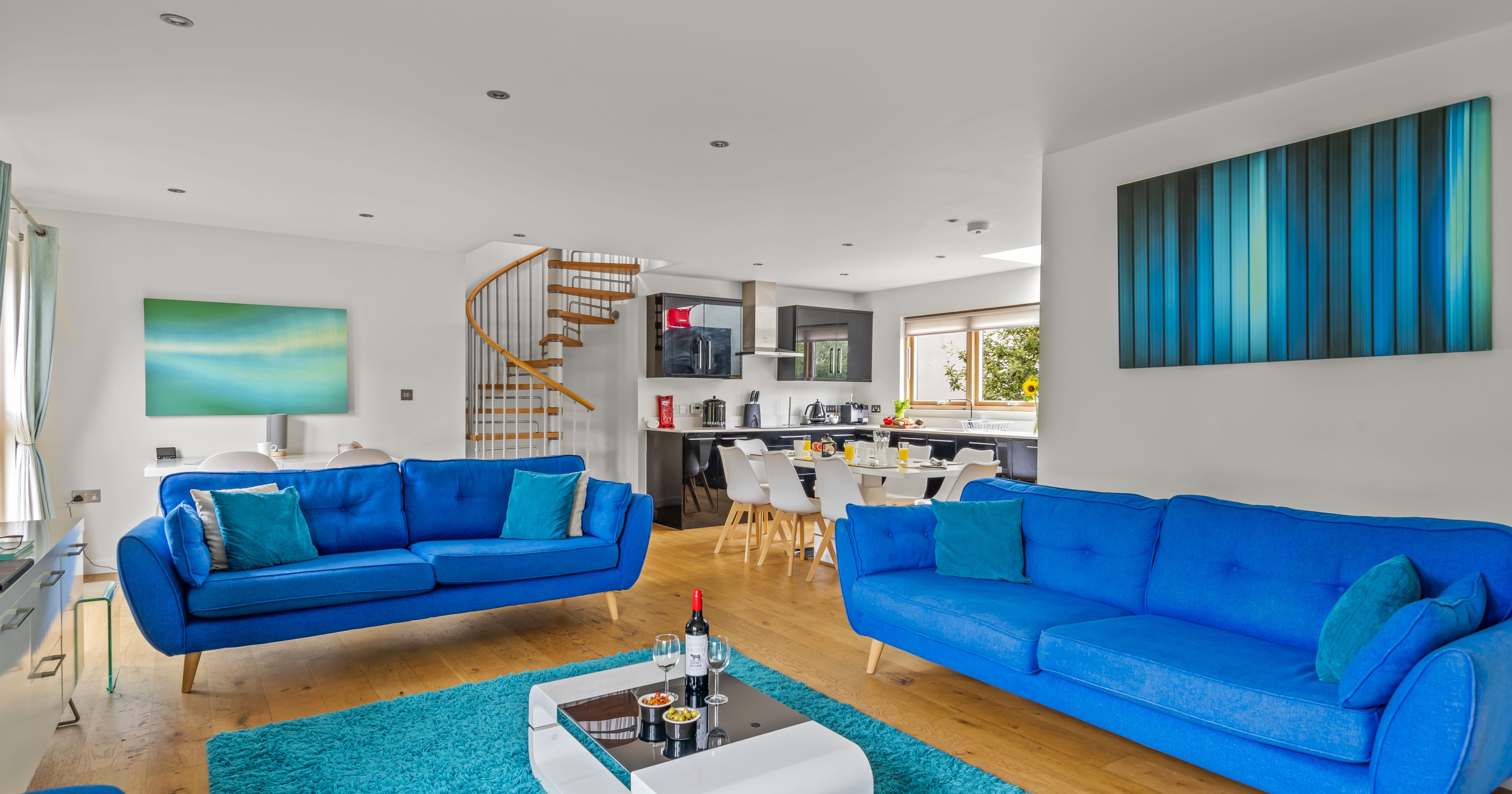 Modern open-plan living room and kitchen with bright blue sofas, abstract wall art, a spiral staircase, and a white coffee table on a blue rug.