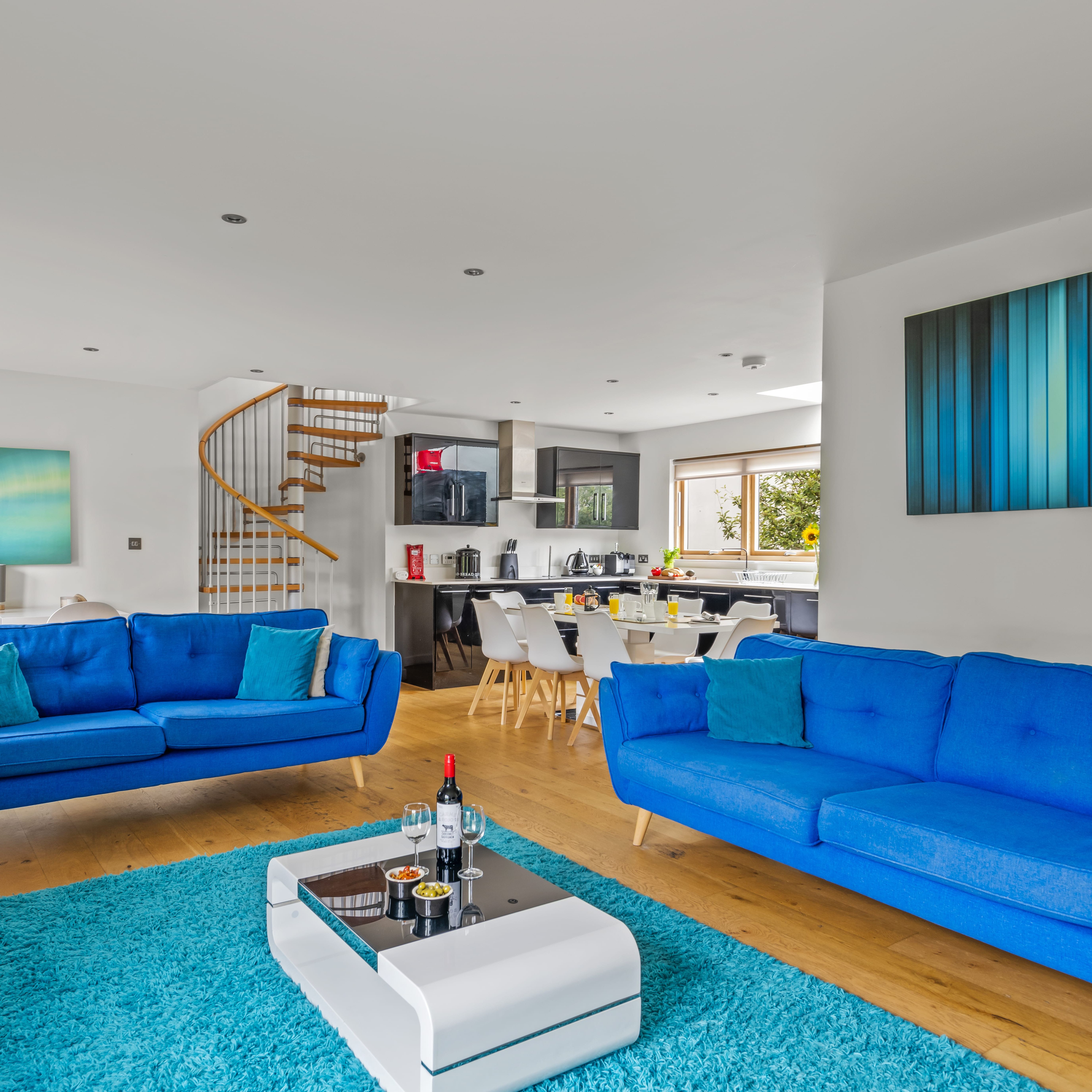Modern open-plan living room and kitchen with bright blue sofas, abstract wall art, a spiral staircase, and a white coffee table on a blue rug.