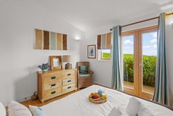 Cozy bedroom with large windows showing a view of green landscape, a bed with breakfast tray, a wooden dresser, and wicker chair.