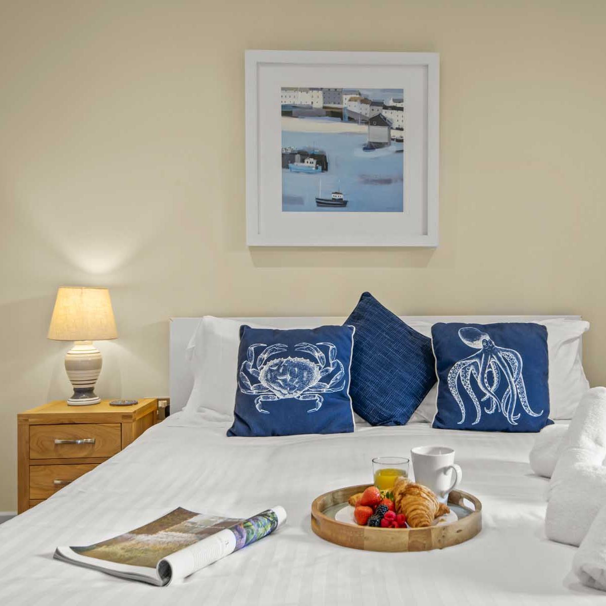 Cozy bedroom with white bedding, blue decorative pillows, and a breakfast tray with pastries, fruit, juice, and coffee on the bed.