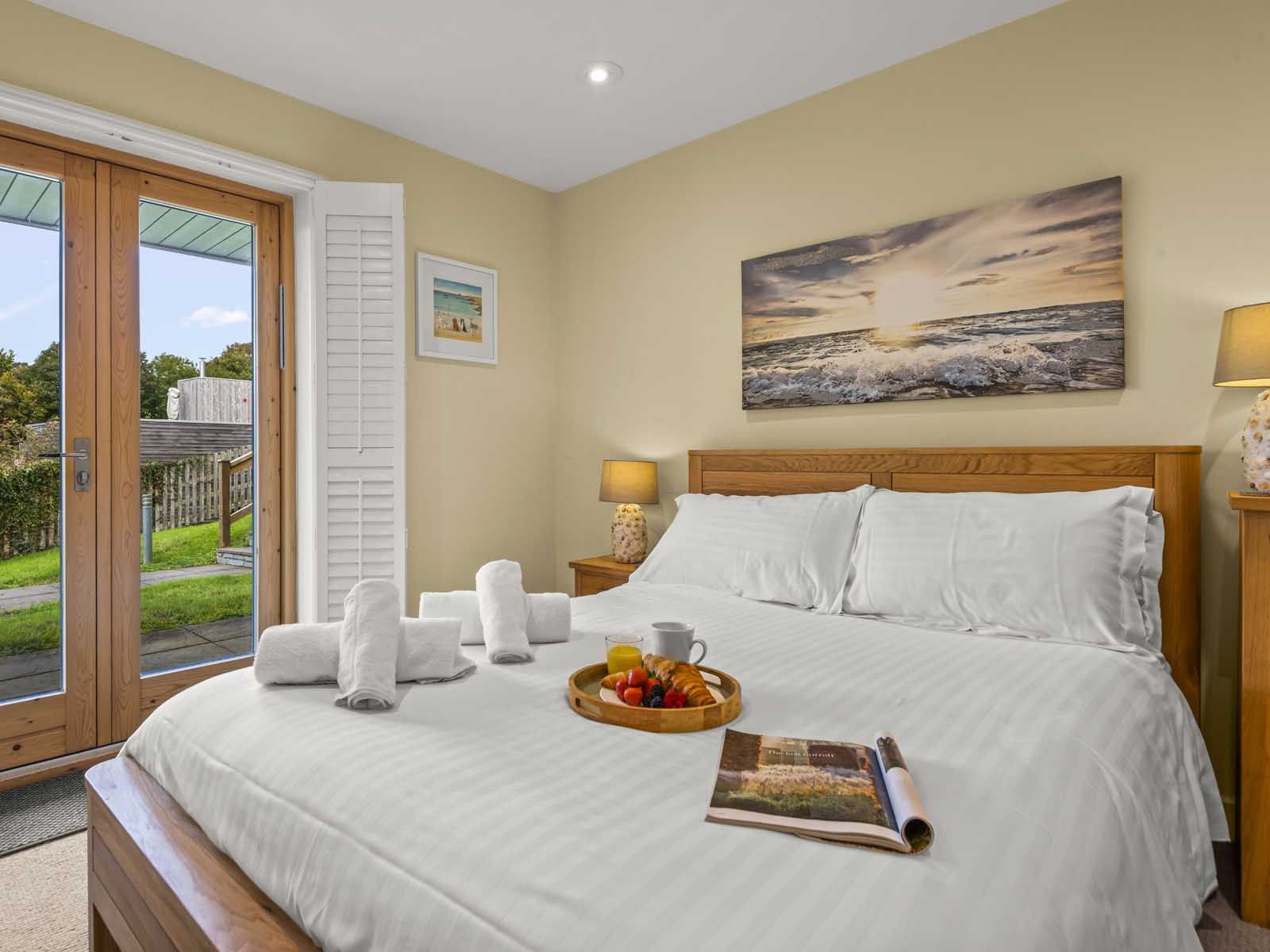 Cozy bedroom with a wooden bed, white linens, a tray with breakfast, rolled towels, and a view to a garden through glass doors.