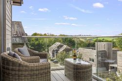 Modern balcony with wicker chairs and glass railing overlooking a countryside view with houses and green hills.