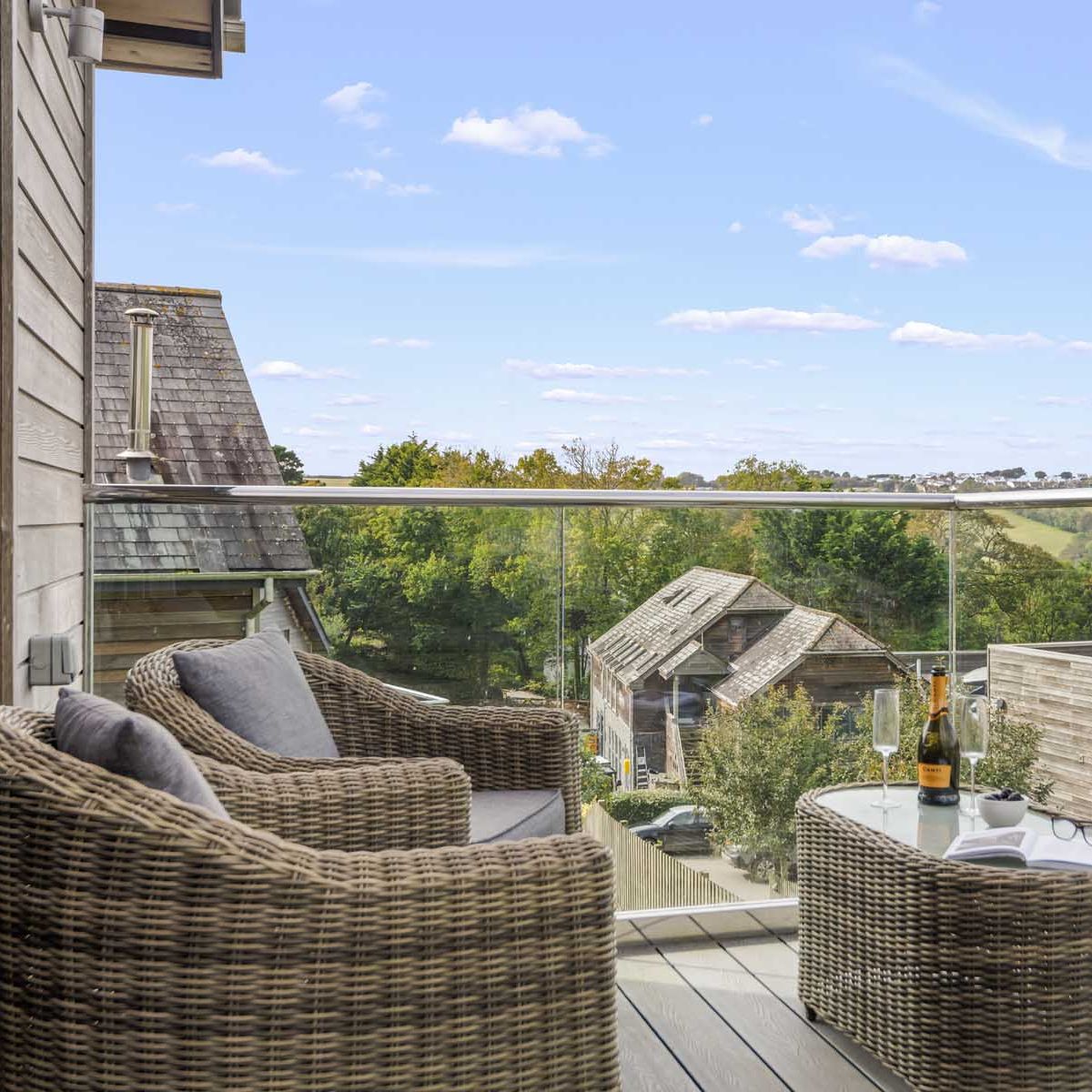 Modern balcony with wicker chairs and glass railing overlooking a countryside view with houses and green hills.