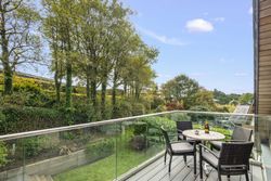 Modern balcony with a glass railing overlooking a lush garden and trees, with a small table and chairs set for drinks.