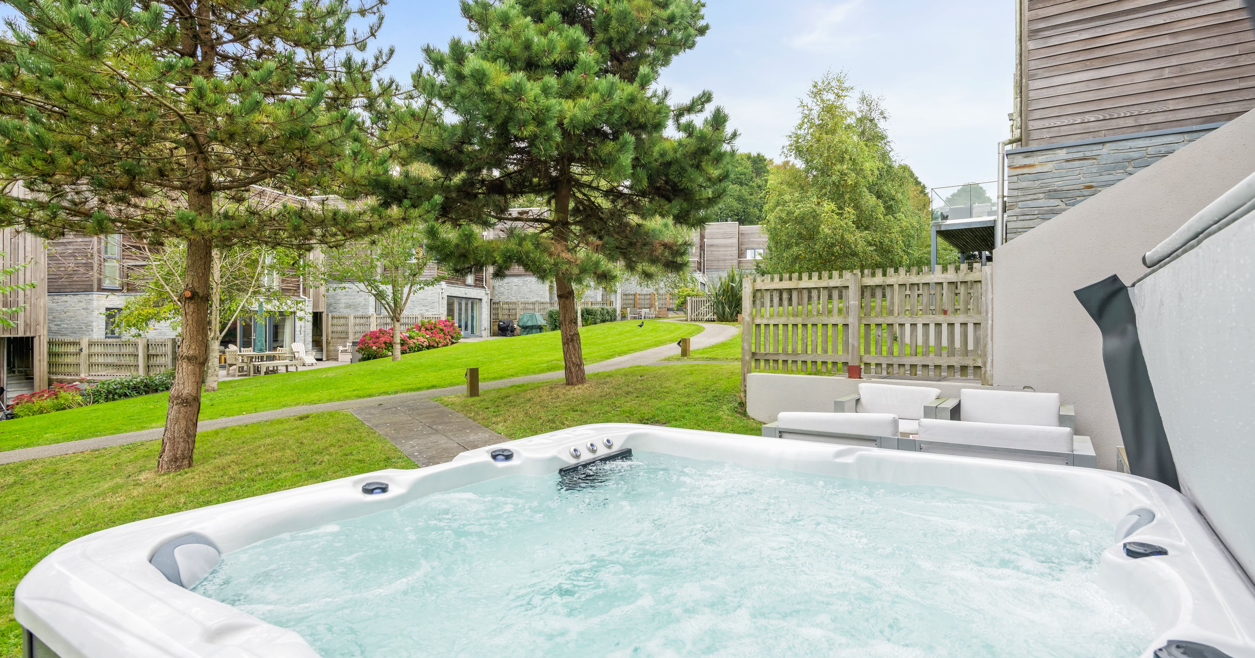 Outdoor hot tub with bubbling water in a garden setting, surrounded by trees, grass, and modern houses.