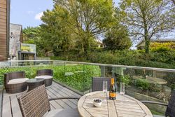 Modern outdoor patio with wicker furniture, glass railing, and a bottle of wine on the table overlooking a lush green garden.
