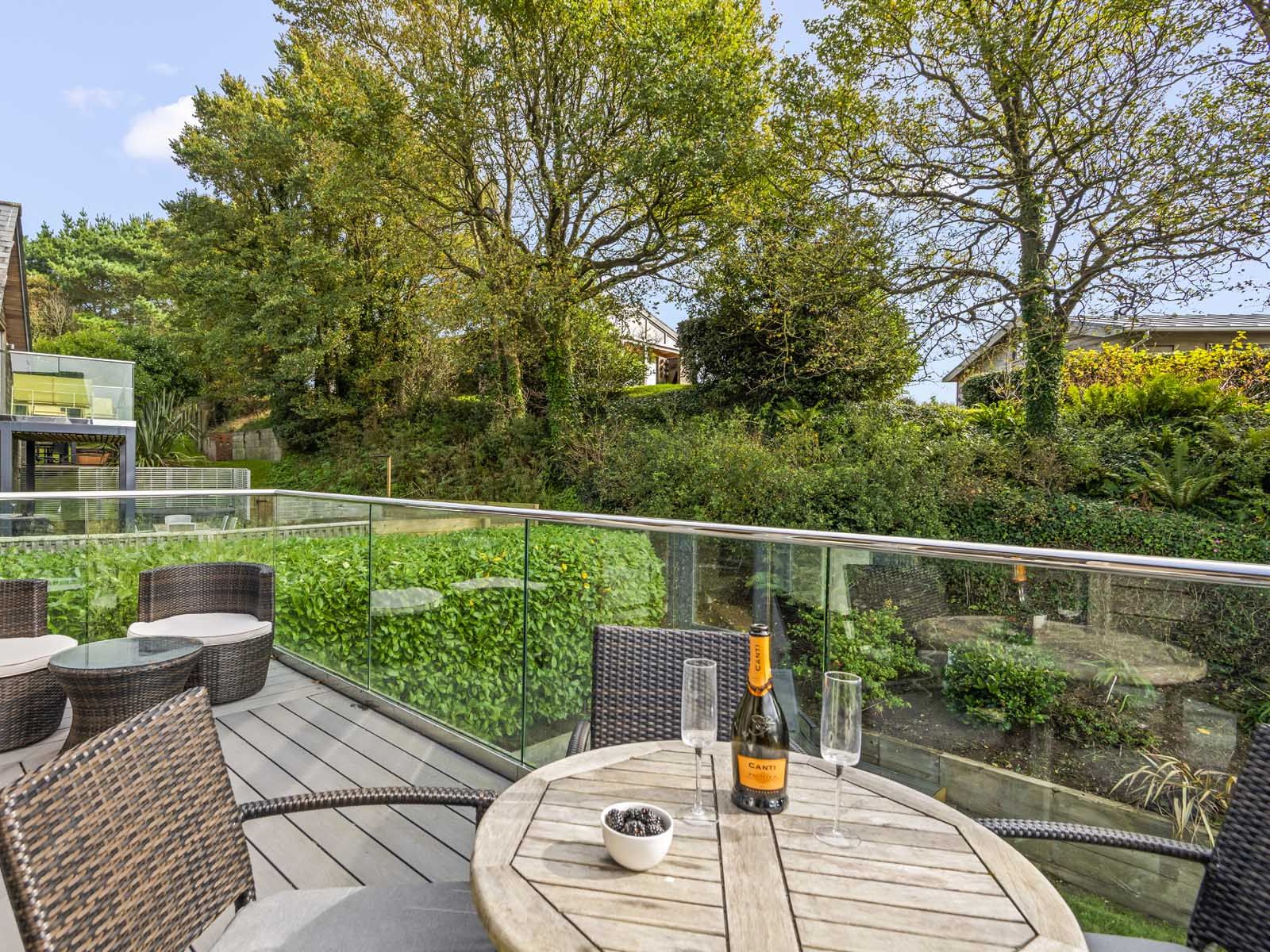 Modern outdoor patio with wicker furniture, glass railing, and a bottle of wine on the table overlooking a lush green garden.