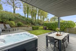 Covered patio with hot tub, outdoor dining table, and lounge chairs overlooking a green garden with trees.