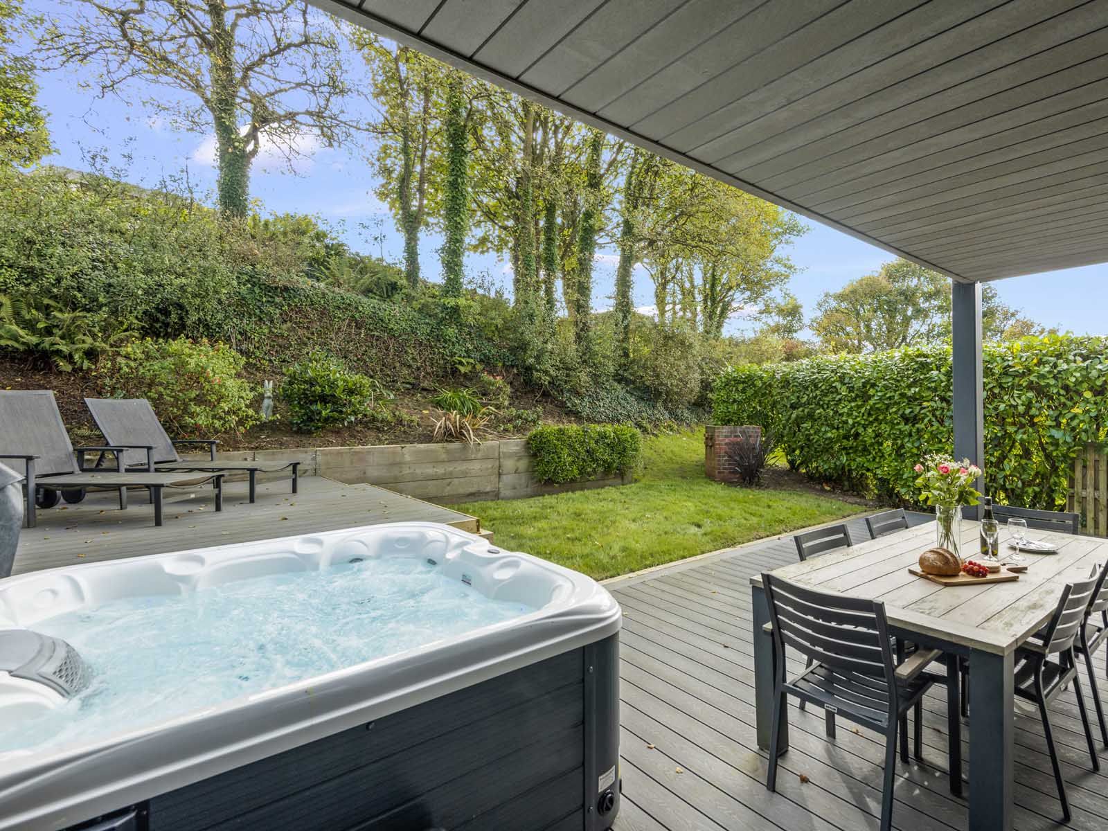 Covered patio with hot tub, outdoor dining table, and lounge chairs overlooking a green garden with trees.