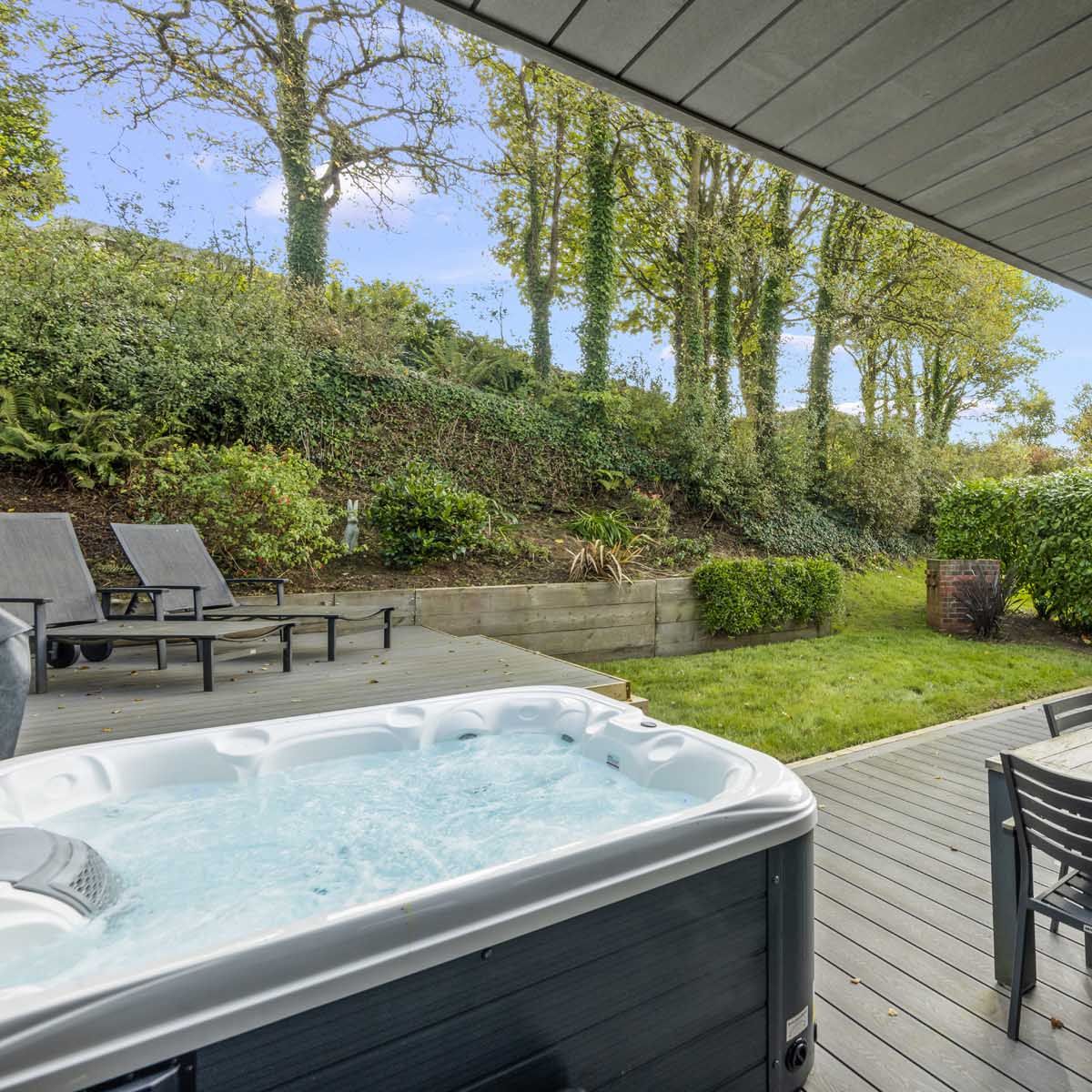 Covered patio with hot tub, outdoor dining table, and lounge chairs overlooking a green garden with trees.