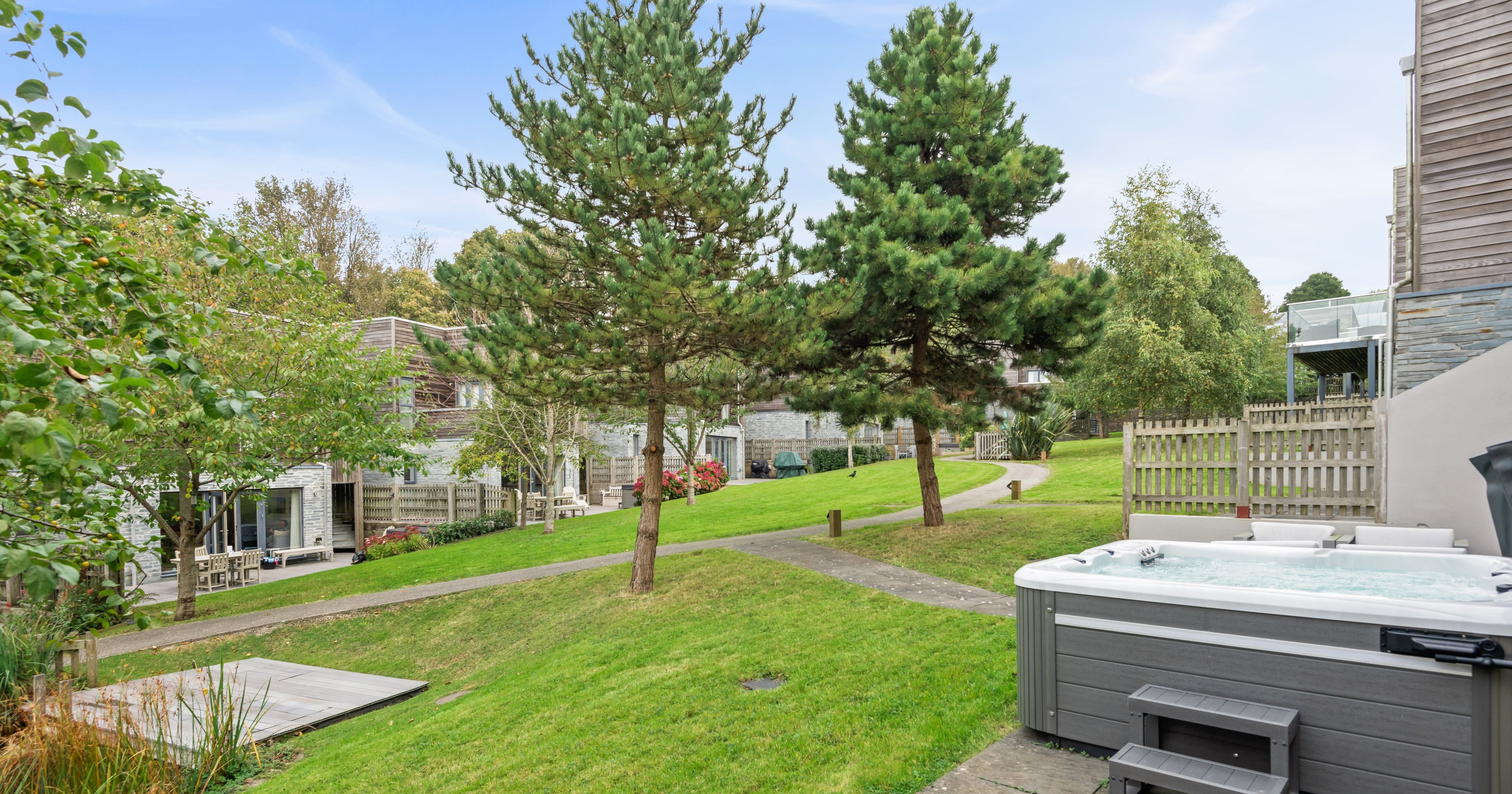 Modern outdoor area with a hot tub, paved patio, green grass, trees, and contemporary buildings in the background under a blue sky.