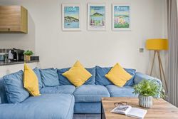 Modern living room with a blue sectional sofa and yellow pillows, wooden coffee table with a plant and glasses, and framed travel posters on the wall.