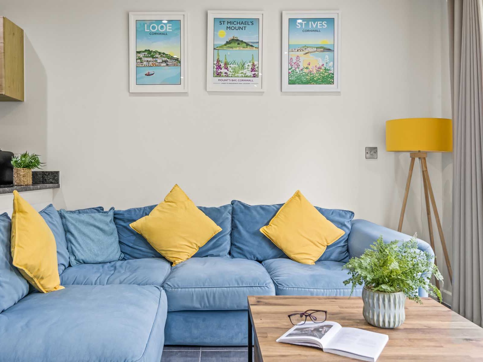 Modern living room with a blue sectional sofa and yellow pillows, wooden coffee table with a plant and glasses, and framed travel posters on the wall.