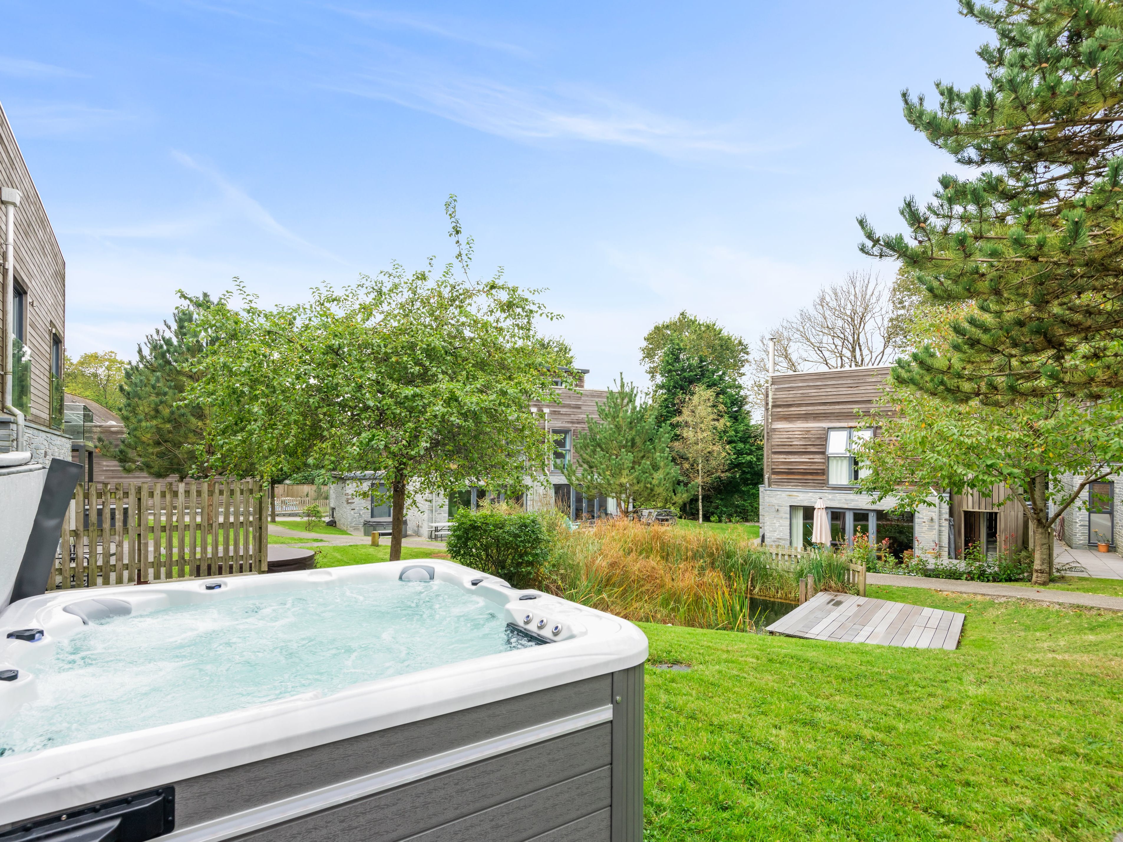 Outdoor hot tub on a grassy lawn with modern wooden houses, trees, and a small pond in the background under a clear blue sky.
