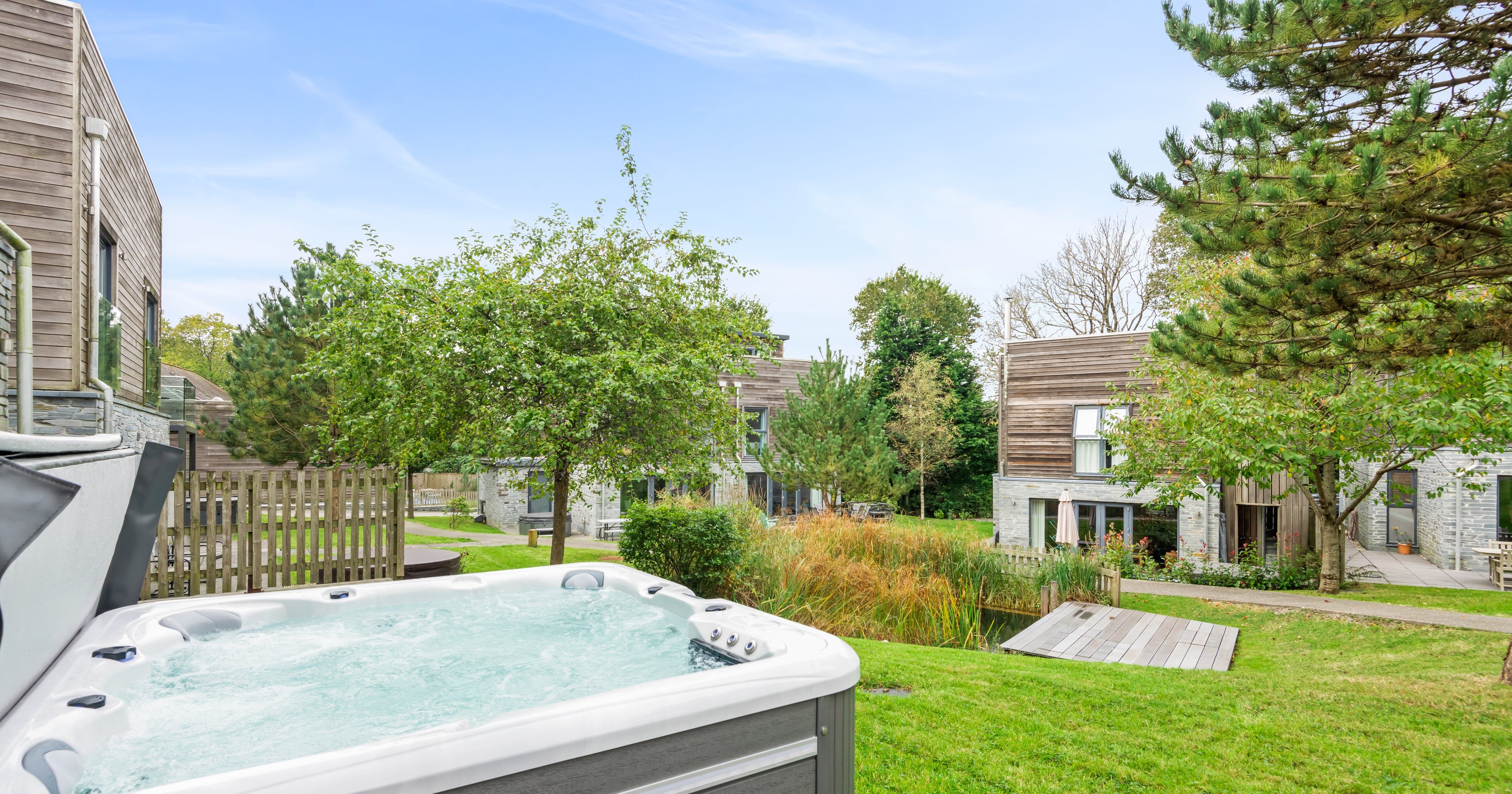 Outdoor hot tub on a grassy lawn with modern wooden houses, trees, and a small pond in the background under a clear blue sky.