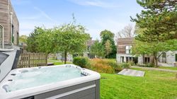 Outdoor hot tub on a grassy lawn with modern wooden houses, trees, and a small pond in the background under a clear blue sky.