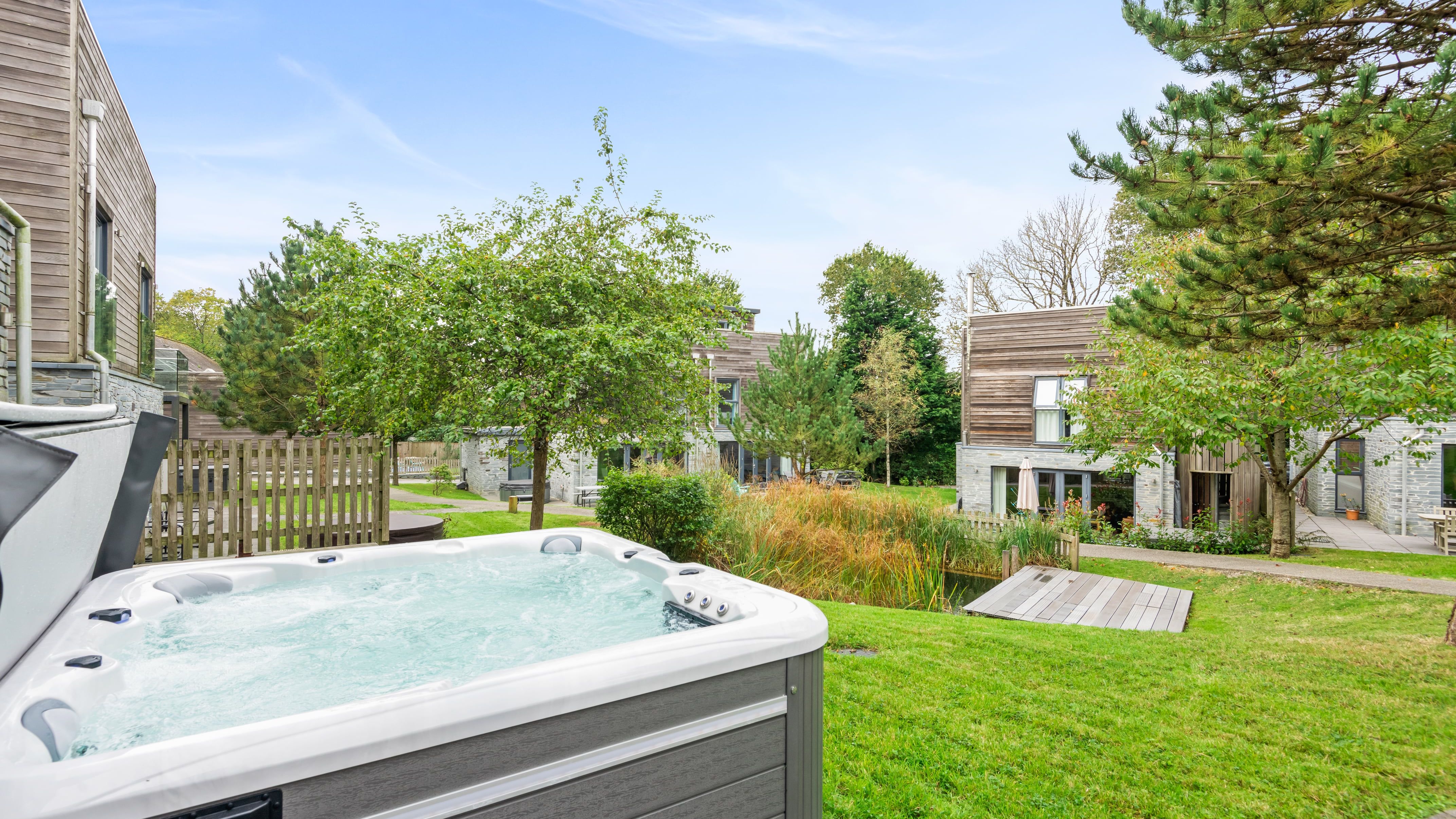 Outdoor hot tub on a grassy lawn with modern wooden houses, trees, and a small pond in the background under a clear blue sky.