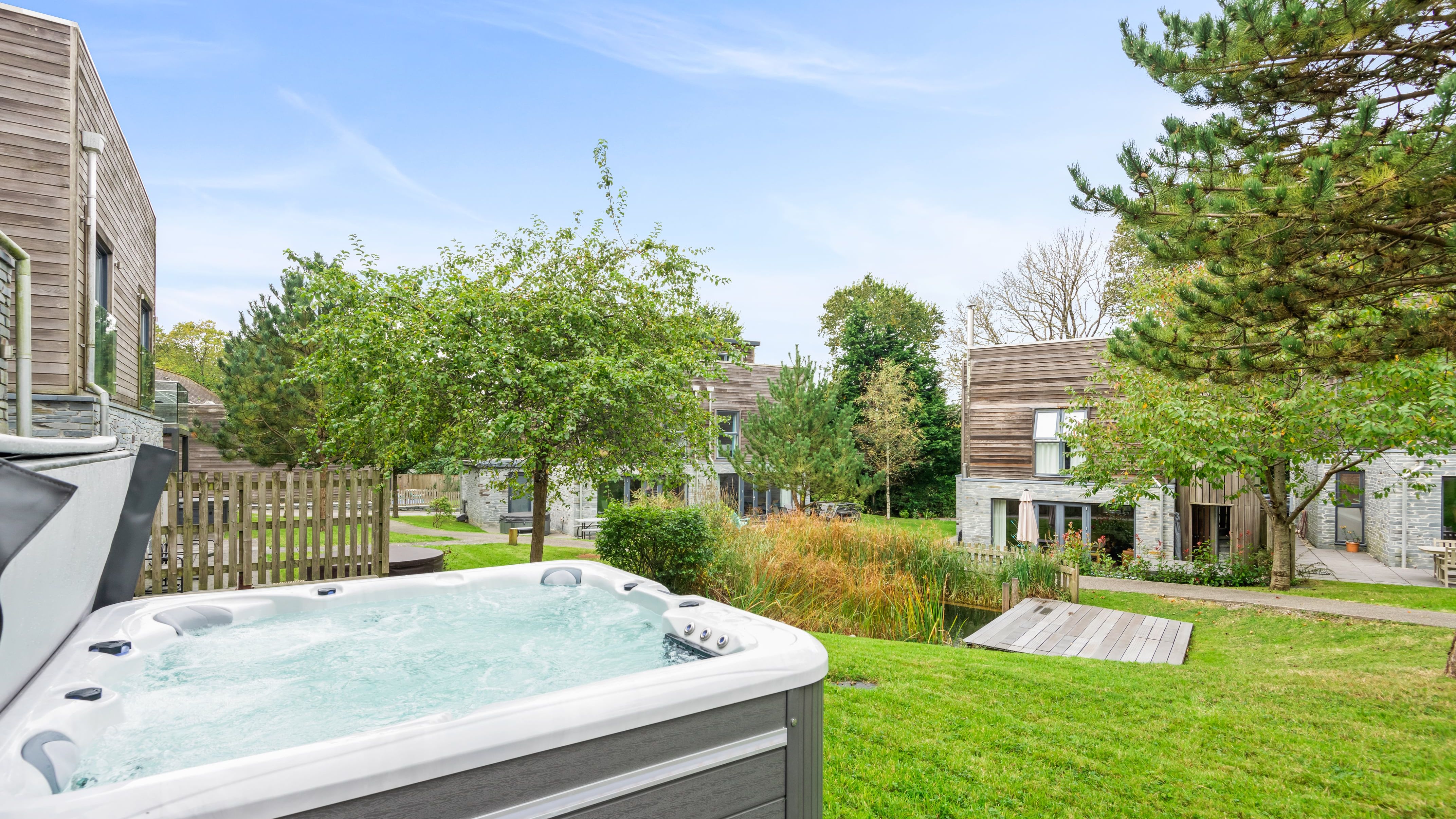 Outdoor hot tub on a grassy lawn with modern wooden houses, trees, and a small pond in the background under a clear blue sky.