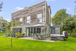 Modern two-story house with wooden and stone exterior, patio furniture, umbrella, and hot tub in a green backyard.