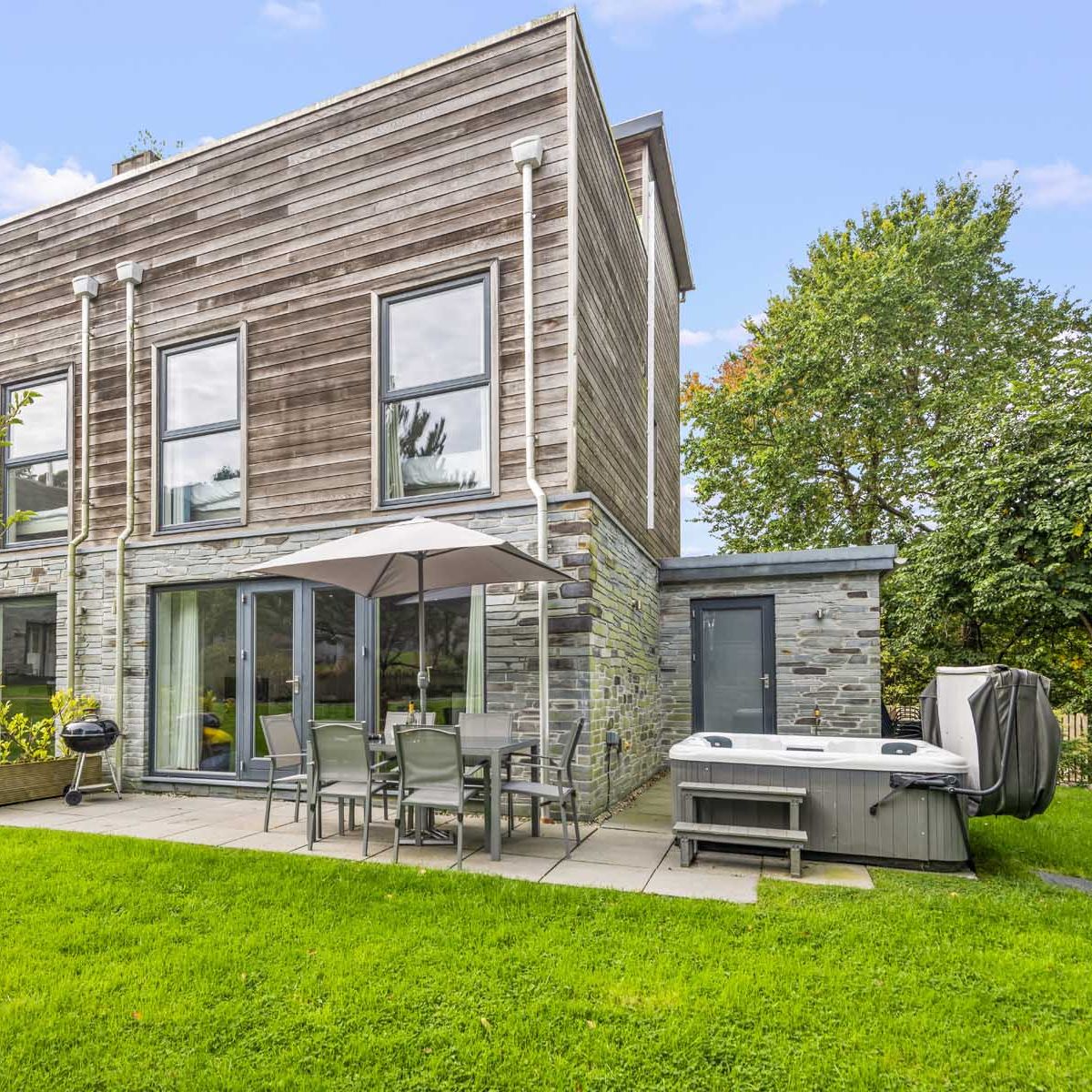 Modern two-story house with wooden and stone exterior, patio furniture, umbrella, and hot tub in a green backyard.