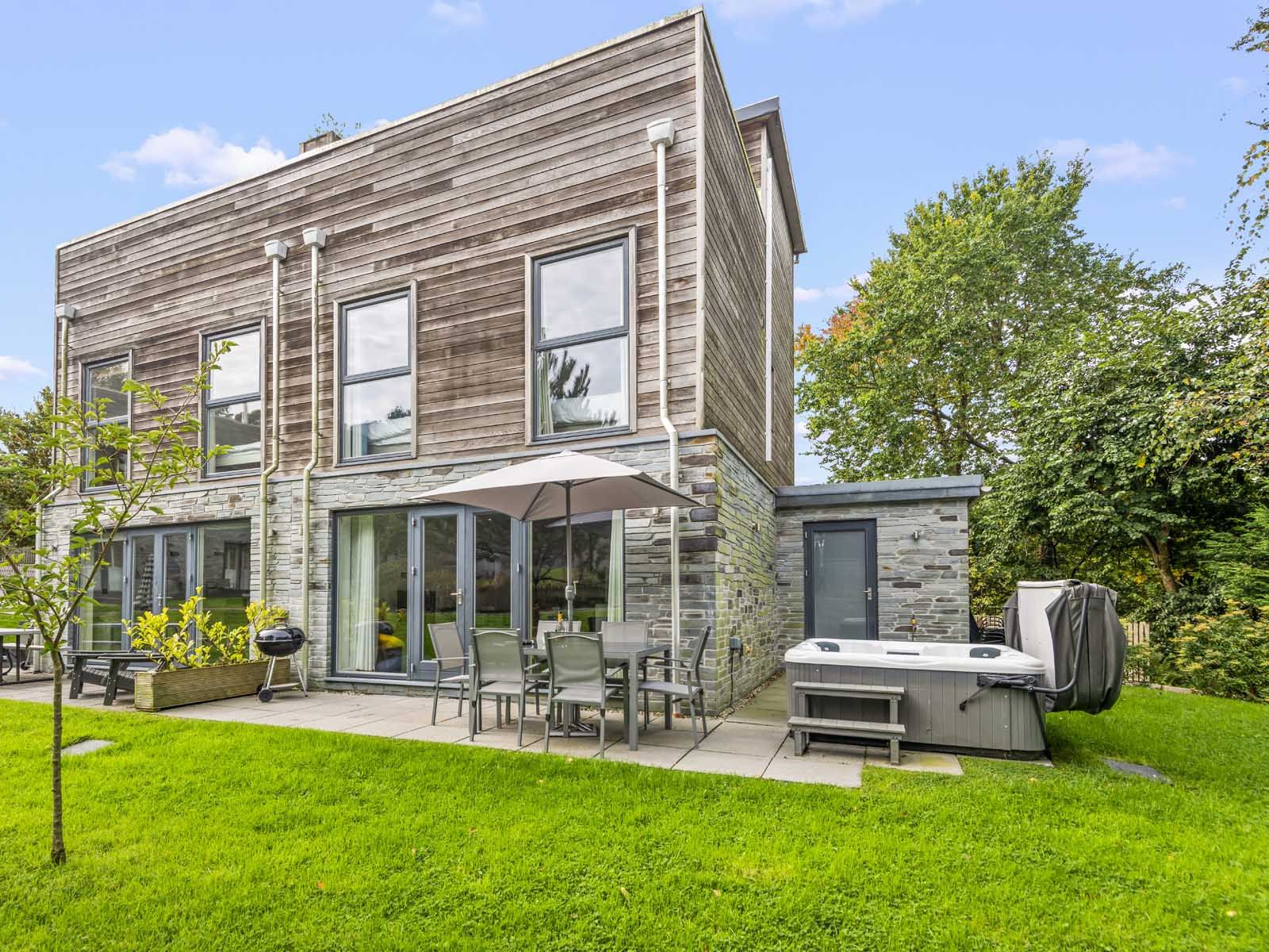 Modern two-story house with wooden and stone exterior, patio furniture, umbrella, and hot tub in a green backyard.